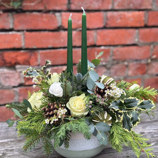 Twelfth Night Christmas table arrangement with green candles ivory roses pine cones waxflower holly and winter foliage displayed in a ceramic bowl.